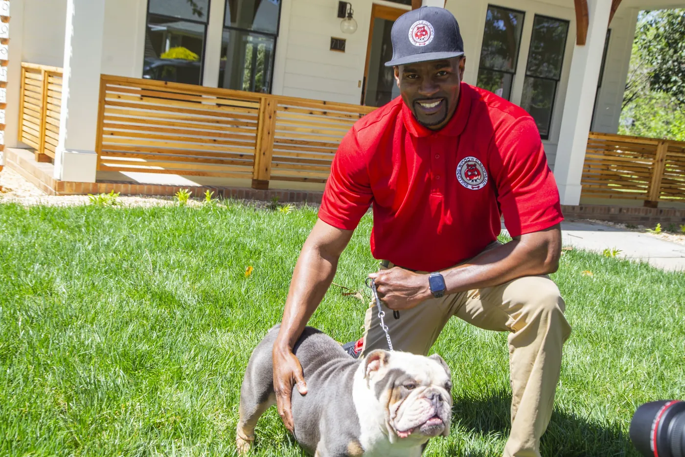 A Bulldog Window Cleaning technician in a red polo and cap kneeling on a lawn next to an English bulldog