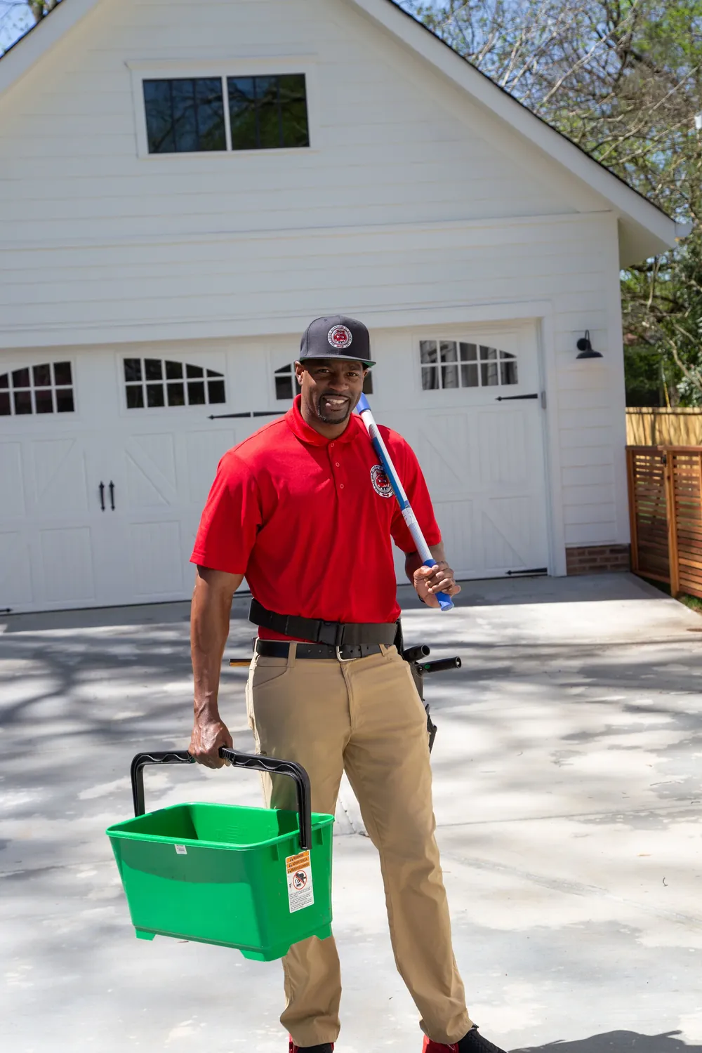 A technician in a red polo holding a pole and a green bucket in front of a white garage