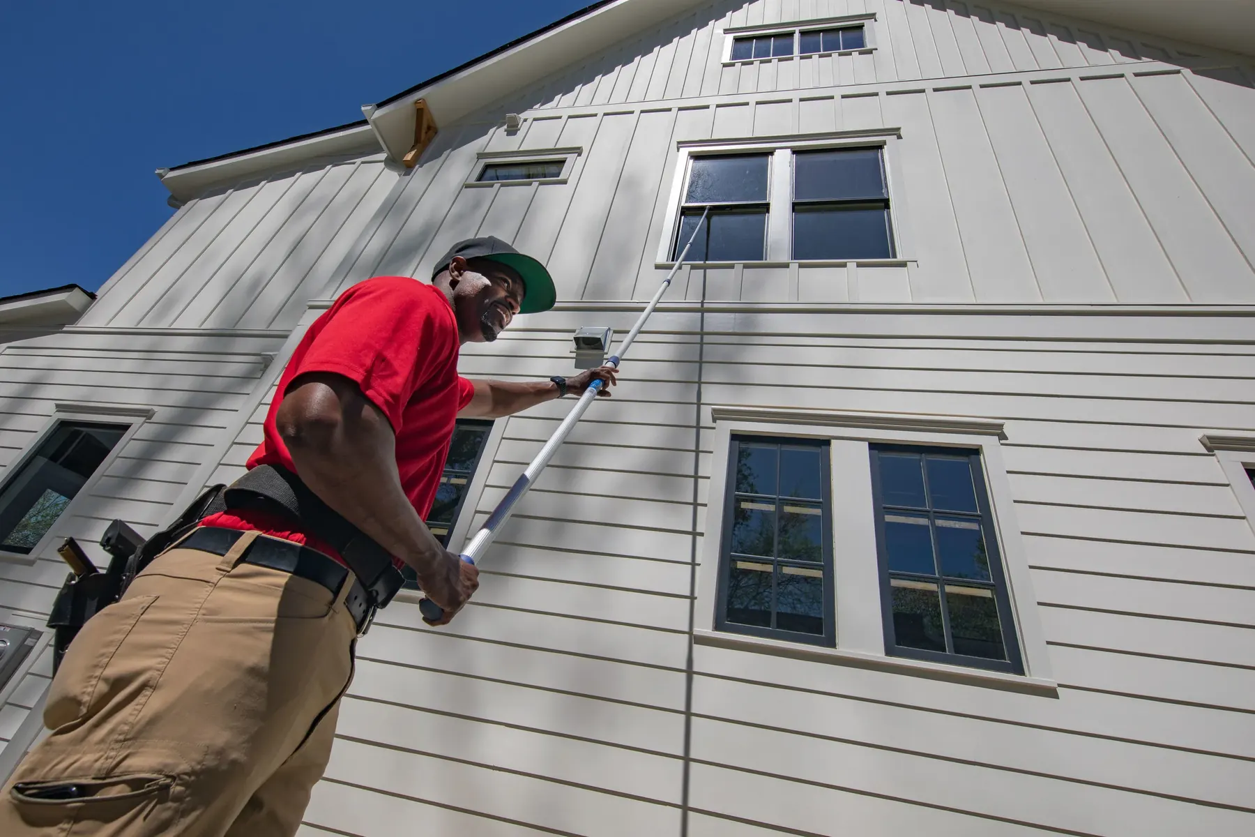 A Bulldog Window Cleaning technician in a red polo using a long pole on the upper windows of a white two-story house
